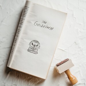 May include: A white book with the title "The Goldfinch" written in black ink. The book has a black and white illustration of a bird with a book in its talons. The book is on a white textured surface.