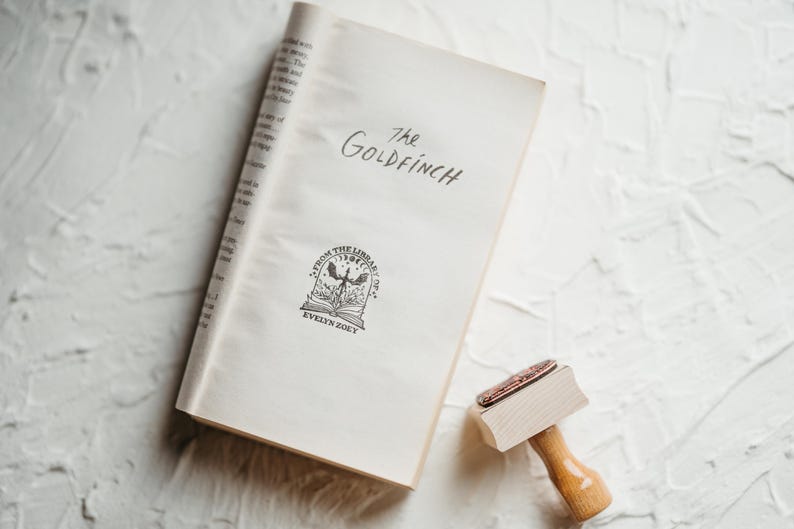 May include: A white book with the title "The Goldfinch" written in black ink. The book has a black and white illustration of a bird with a book in its talons. The book is on a white textured surface.
