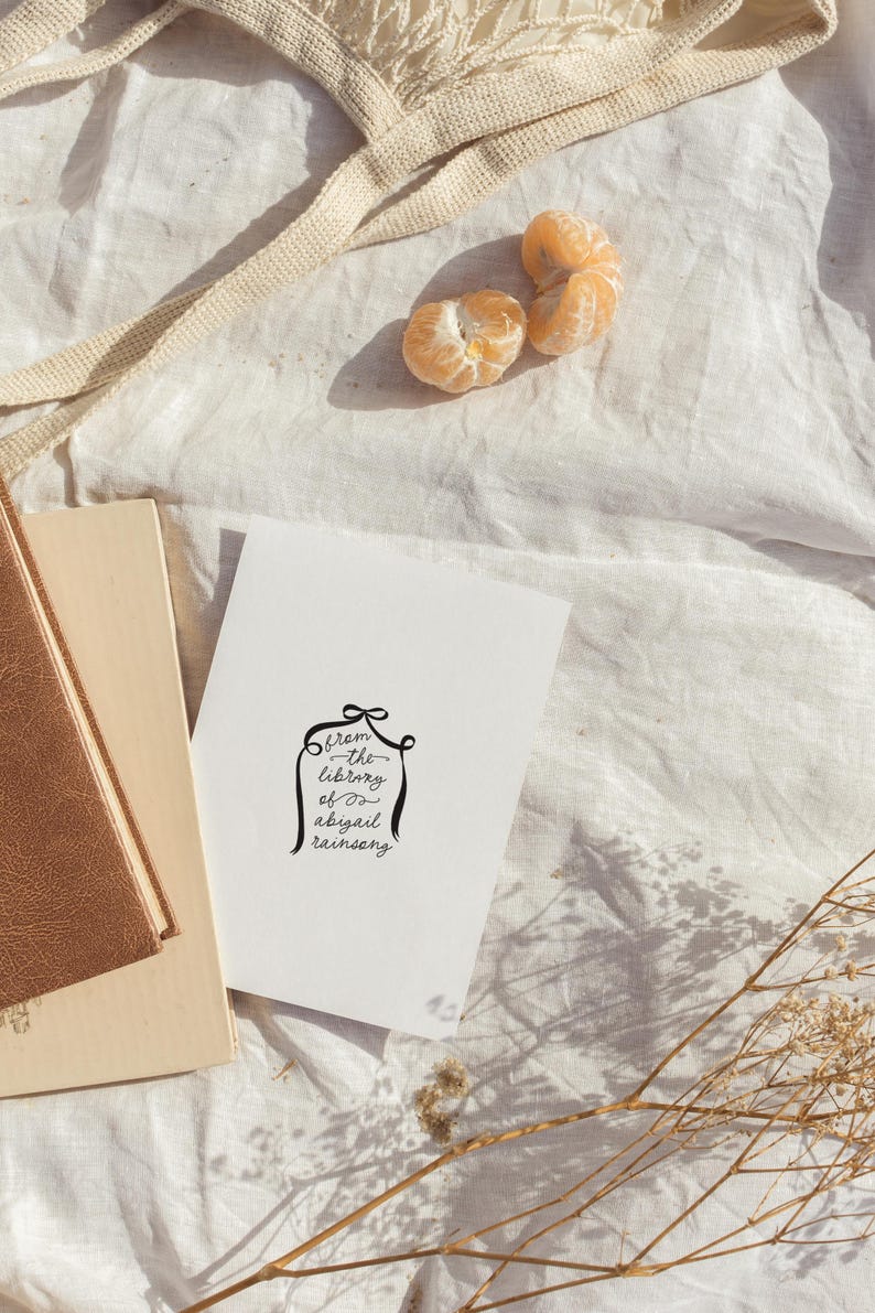 Peut inclure: Une carte blanche avec une calligraphie noire qui dit "from the library of abigail rainsong". La carte est sur une surface en tissu blanc avec un livre brun et quelques fleurs s&eacute;ch&eacute;es.