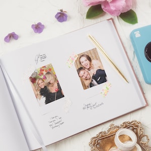 May include: Open photo album with two photos, signatures, and a gold pen. A light blue Kodak camera and decorative items are on the white surface. The album has a pink cover and a white ribbon.