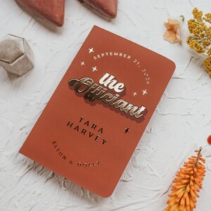 May include: A terracotta-colored wedding program with gold foil lettering that reads "the Officiant." The program includes the date, names, and is surrounded by decorative elements like flowers and a ring box.
