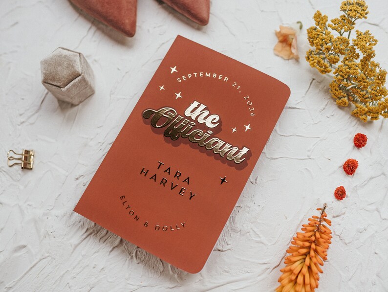 May include: A terracotta-colored wedding program with gold foil lettering that reads "the Officiant." The program includes the date, names, and is surrounded by decorative elements like flowers and a ring box.