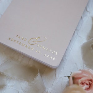 K&ouml;nnte beinhalten: Ein hellrosa Hochzeitsnotizbuch mit goldfarbenen Buchstaben. Der Text lautet "JANE & TIMOTHY" und "SEPTEMBER 21, 2026". Das Notizbuch ist von weichem Stoff und rosa Rosen umgeben, was auf ein Hochzeitsthema hindeutet.