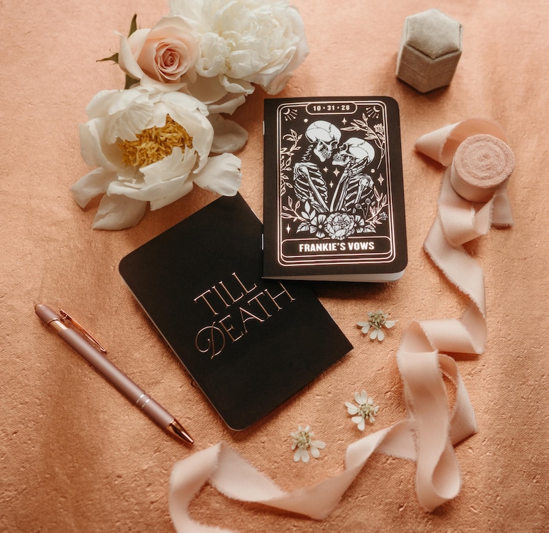 May include: A flat lay featuring wedding stationery and floral accents. Two black notebooks with gold lettering and a pen are displayed alongside a ring box, ribbon, and flowers. One notebook reads "Till Death" and the other "Frankie's Vows" with a skeleton couple illustration.
