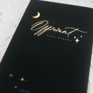 May include: Black notebook with gold foil lettering reading "Officiant TARA HARVEY". The design includes a crescent moon and stars. The notebook is on a white surface.