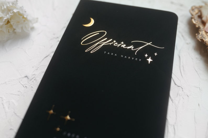 May include: Black notebook with gold foil lettering reading "Officiant TARA HARVEY". The design includes a crescent moon and stars. The notebook is on a white surface.