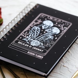 Puede incluir: Agenda de bodas negra con espiral con una ilustraci&oacute;n decorativa de esqueletos y elementos florales. La portada dice "DELILAH & JOHNNY WEDDING PLANNER". Un bol&iacute;grafo dorado y rosas rojas est&aacute;n en el fondo.