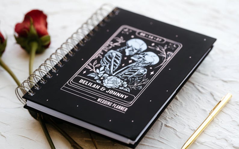 Puede incluir: Agenda de bodas negra con espiral con una ilustraci&oacute;n decorativa de esqueletos y elementos florales. La portada dice "DELILAH & JOHNNY WEDDING PLANNER". Un bol&iacute;grafo dorado y rosas rojas est&aacute;n en el fondo.