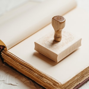 Op de afbeelding: Een houten stempel met de tekst "TA' AMELIO" en een sierlijk ontwerp ligt op een lichtgekleurd boek. De stempel heeft een afgeronde handgreep. Het boek is open en toont verouderde, cr&egrave;mekleurige pagina's.