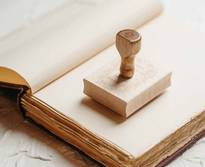 Op de afbeelding: Een houten stempel met de tekst "TA' AMELIO" en een sierlijk ontwerp ligt op een lichtgekleurd boek. De stempel heeft een afgeronde handgreep. Het boek is open en toont verouderde, cr&egrave;mekleurige pagina's.