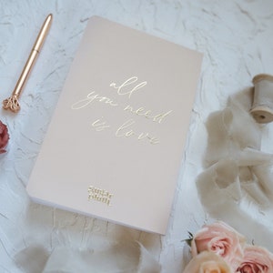 K&ouml;nnte beinhalten: Ein rosafarbenes Notizbuch mit dem goldfarbenen Text "all you need is love". Ein goldener Stift und Rosen liegen neben dem Notizbuch. Das Sugar Plum-Logo befindet sich am unteren Rand des Notizbuchs. Der Hintergrund ist eine strukturierte wei&szlig;e Oberfl&auml;che.