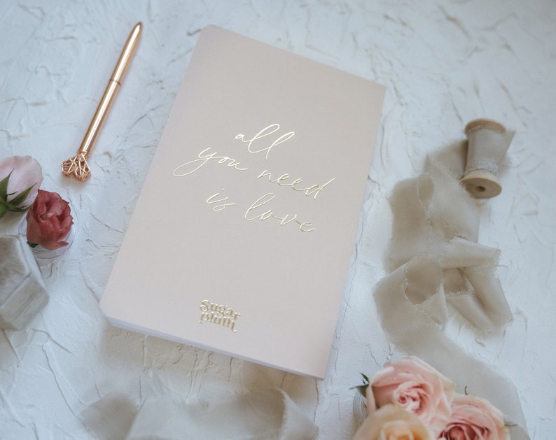 K&ouml;nnte beinhalten: Ein rosafarbenes Notizbuch mit dem goldfarbenen Text "all you need is love". Ein goldener Stift und Rosen liegen neben dem Notizbuch. Das Sugar Plum-Logo befindet sich am unteren Rand des Notizbuchs. Der Hintergrund ist eine strukturierte wei&szlig;e Oberfl&auml;che.