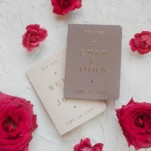 May include: Two ivory wedding vow books with gold lettering. The top book has the text "his vows" and the bottom book has the text "her vows". Both books have the names "Ruby & John" and the date "Sept. 21, 2023".