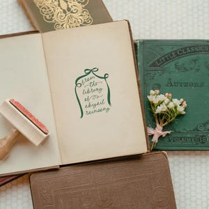 Peut inclure: Un tampon vert avec le texte "from the library of abigail rainsong" sur fond blanc. Le tampon est sur un livre brun avec un tampon en caoutchouc en bois &agrave; c&ocirc;t&eacute;. Il y a un livre vert avec le texte "Little Classics" sur la couverture.