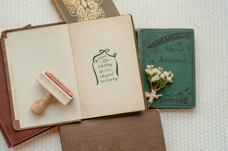 Peut inclure: Un tampon vert avec le texte "from the library of abigail rainsong" sur fond blanc. Le tampon est sur un livre brun avec un tampon en caoutchouc en bois &agrave; c&ocirc;t&eacute;. Il y a un livre vert avec le texte "Little Classics" sur la couverture.