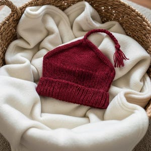 May include: A knitted burgundy baby hat with a tassel, resting in a woven basket with a white blanket. The hat has a folded brim and a pointed top. The basket is filled with a soft, white blanket.