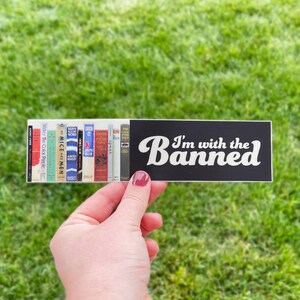 May include: A rectangular sticker featuring a row of colorful book spines on the left side and the white text "I'm with the Banned" on a black background on the right side. The sticker displays various book titles including Of Mice and Men.