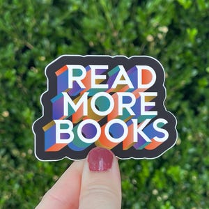 May include: A die-cut sticker with a black border featuring the white text READ MORE BOOKS in a bold, blocky font. The letters have a three-dimensional effect with a colorful gradient pattern of blue, orange, and green layered behind the white text.