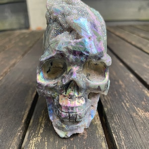 Puede incluir: Un cráneo de cristal hecho de piedra gris y negra iridiscente y brillante. El cráneo está detallado con cuencas de los ojos, una nariz y dientes.