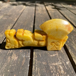 May include: A carved, cream-colored sculpture with a stylized, abstract design. The sculpture has a rounded top and a rectangular base with carved details. The piece is displayed on a weathered wooden surface.