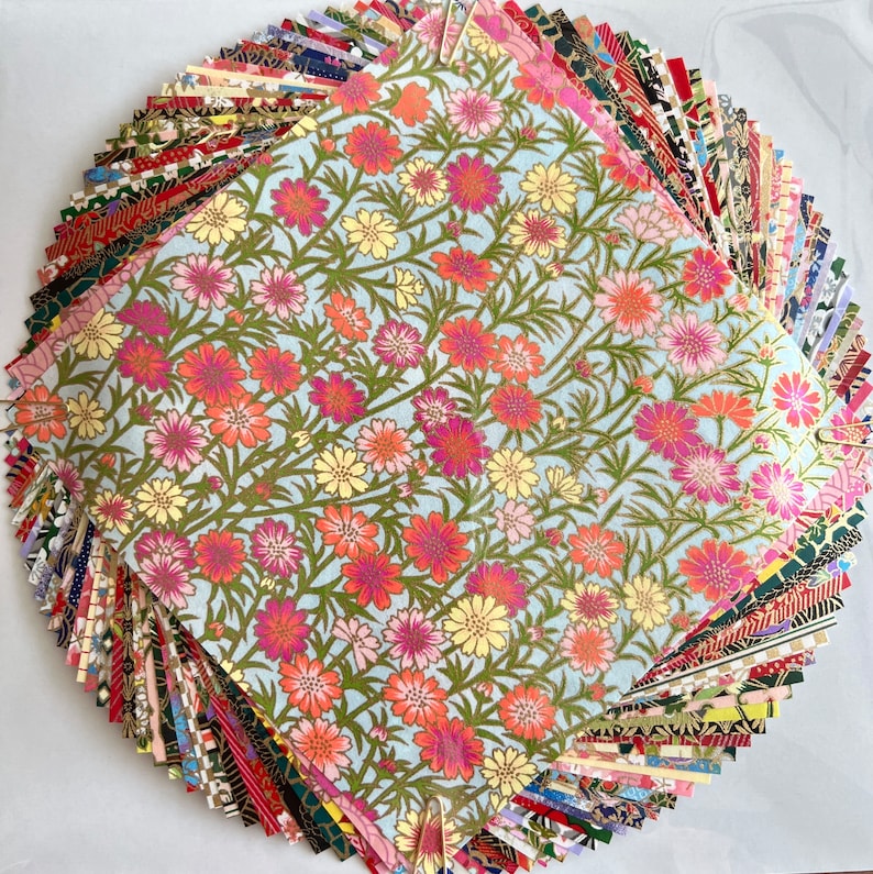Peut inclure: Une pile de papier origami &agrave; motifs color&eacute;s, avec une seule feuille de papier bleu clair avec un motif floral sur le dessus. Le motif floral pr&eacute;sente des fleurs roses, rouges et jaunes sur un fond bleu clair.