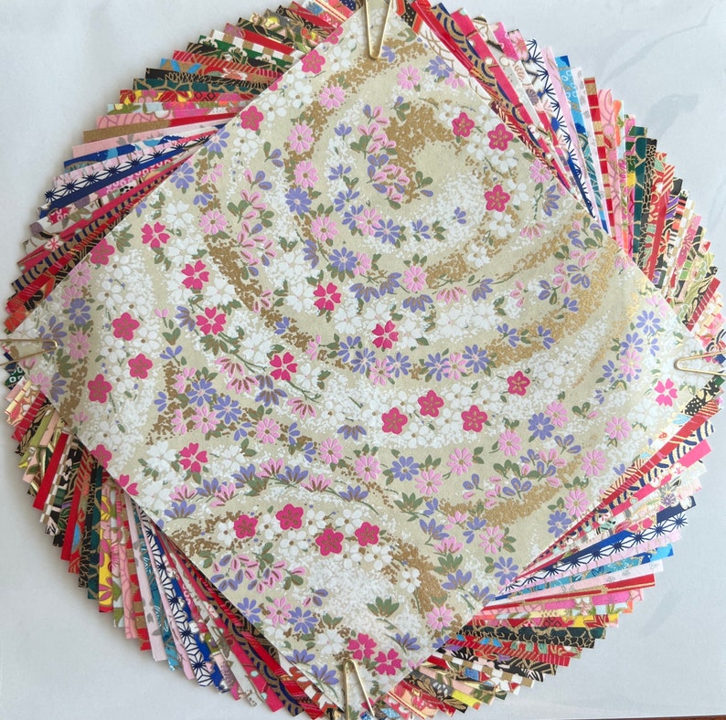 Peut inclure: Un &eacute;ventail de carr&eacute;s de papier aux motifs color&eacute;s, pli&eacute;s et &eacute;vas&eacute;s, avec un seul carr&eacute; au centre pr&eacute;sentant un fond blanc avec un motif floral d&eacute;licat en rose, violet et or.