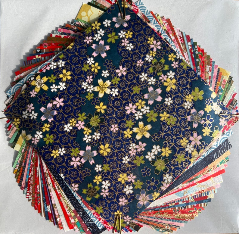 Peut inclure: Un &eacute;ventail de carr&eacute;s de papier aux motifs color&eacute;s, avec un carr&eacute; bleu fonc&eacute; avec un motif floral blanc et or au centre.