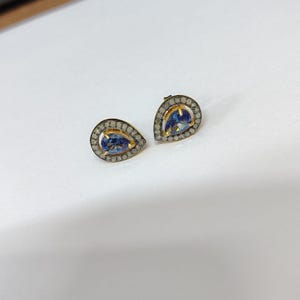 Pode incluir: Um par de brincos em forma de gota. Cada brinco apresenta um centro de pedra preciosa azul vibrante, rodeado por um halo de pedras menores e transparentes. A cravação é dourada, criando um contraste marcante.