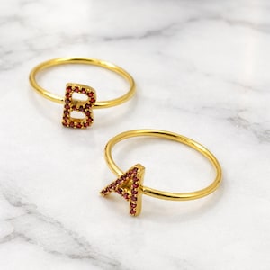 May include: Two gold-colored rings, one with the letter "B" and the other with the letter "A". The letters are encrusted with small, red gemstones. The rings are displayed on a white and gray marble surface.