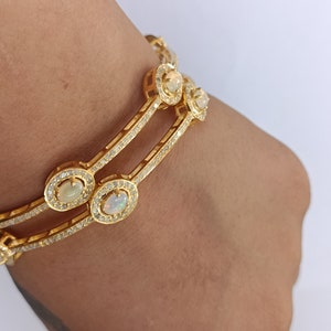 May include: A gold bangle bracelet with a double row of sparkling stones and oval-shaped opals. The bracelet is on a person's wrist.
