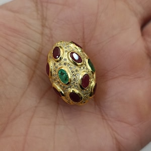 Könnte beinhalten: Ein ovaler Anhänger aus Gold mit roten und grünen Edelsteinen, die in einen goldenen Rahmen gefasst sind. Der Anhänger ist von kleineren Diamanten umgeben.