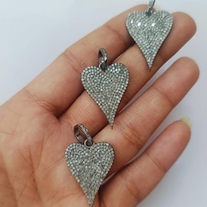 May include: Three heart-shaped pendants, each covered in small, sparkling stones. The pendants have a silver-colored bail at the top for hanging. The heart design is elongated with a pointed bottom. The pendants are displayed against a neutral background.