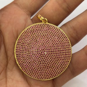 Puede incluir: Un colgante de oro con un diseño redondo y plano. El colgante está cubierto de pequeñas piedras preciosas rosas.