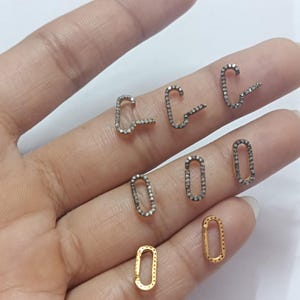 Könnte beinhalten: Eine Sammlung kleiner, ovaler und C-förmiger Anhänger aus Silber und Gold. Jeder Anhänger ist mit winzigen, funkelnden Diamanten verziert.