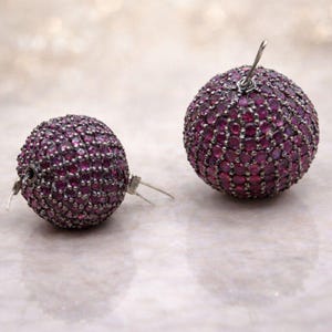 Pode incluir: Um par de brincos redondos. Cada brinco é uma esfera coberta por pequenas pedras preciosas rosa escuro. Os brincos têm um pino e um fecho prateados. O fundo é de uma cor neutra suave e desfocada.