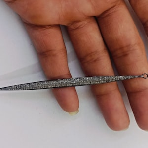 May include: A long, slender, needle-like jewellery component featuring a dark, oxidised metal finish encrusted with numerous small, sparkling pav&eacute;-set stones. The piece tapers to a sharp point at one end and features a small circular loop at the other.