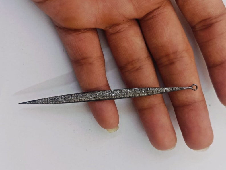 May include: A long, slender, needle-like jewellery component featuring a dark, oxidised metal finish encrusted with numerous small, sparkling pav&eacute;-set stones. The piece tapers to a sharp point at one end and features a small circular loop at the other.