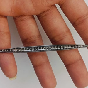 May include: A long, slender, silver-toned metal tool with a pointed tip on one end and a small circular loop on the other. The central body of the tool features a textured, cross-hatched pattern along its length, creating a distinct grid-like appearance.