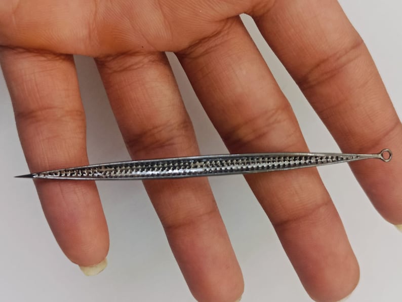 May include: A long, slender, silver-toned metal tool with a pointed tip on one end and a small circular loop on the other. The central body of the tool features a textured, cross-hatched pattern along its length, creating a distinct grid-like appearance.