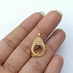May include: Gold-tone clasp with clear cubic zirconia stones. The clasp is shaped like a teardrop and has a round opening.