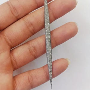 May include: A long, slender, needle-shaped pendant featuring a dark metallic frame encrusted with numerous small, sparkling white stones. The piece tapers to a sharp point at the bottom and includes a small circular loop at the top for attachment.