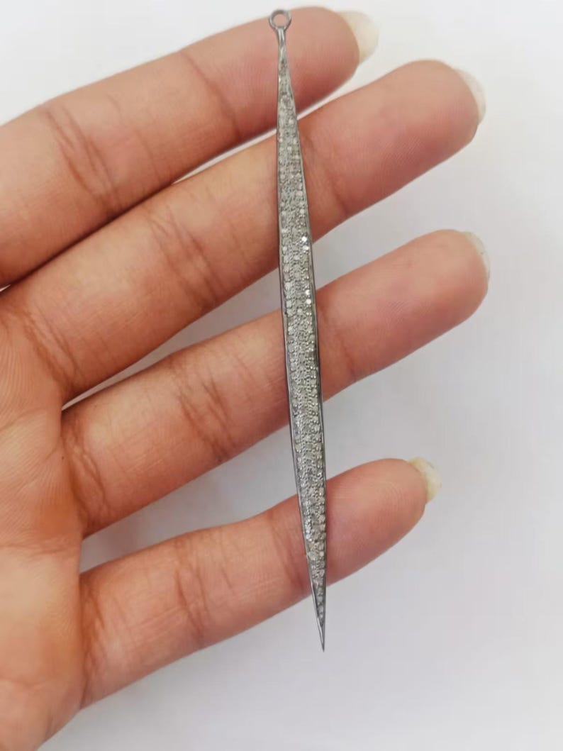 May include: A long, slender, needle-shaped pendant featuring a dark metallic frame encrusted with numerous small, sparkling white stones. The piece tapers to a sharp point at the bottom and includes a small circular loop at the top for attachment.
