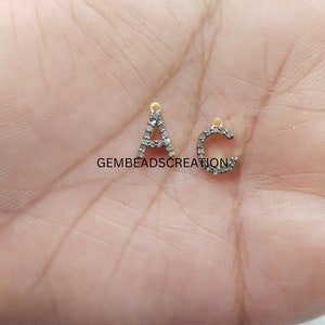 May include: Two small, silver-coloured letter charms, an "A" and a "C", are displayed. The letters are encrusted with small, dark stones and have gold-coloured loops at the top for hanging. The letters are on a light background.