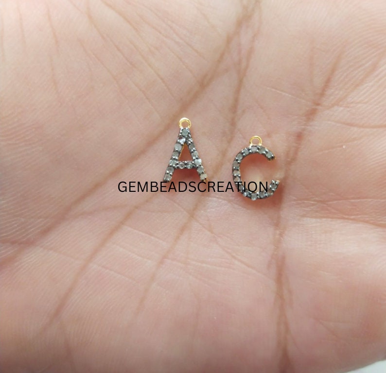 May include: Two small, silver-coloured letter charms, an "A" and a "C", are displayed. The letters are encrusted with small, dark stones and have gold-coloured loops at the top for hanging. The letters are on a light background.