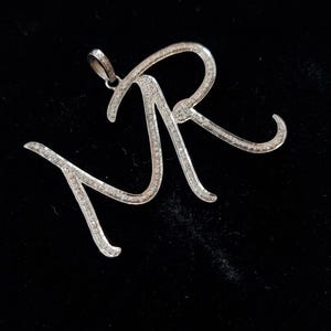 May include: A silver-toned metal pendant shaped as the cursive letters M and R, featuring a textured surface encrusted with numerous small, clear, sparkling stones. The pendant includes a small circular bail at the top for attachment to a chain.