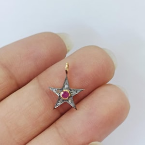Pode incluir: Um amuleto de estrela de prata com uma pedra vermelha no centro e pequenas pedras transparentes ao redor da borda. O amuleto está em uma corrente de ouro.