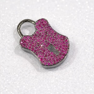 May include: A silver-toned padlock pendant with a keyhole design. The padlock is encrusted with small, vibrant pink gemstones. The pendant has a curved top and a rounded bottom, set against a white, glittery background.