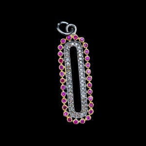 Puede incluir: Un colgante de plata con un diseño ovalado alargado. El colgante está delineado con pequeñas gemas rosas vibrantes y presenta una fila central de gemas claras y marrones. La argolla es plateada. El fondo es negro.