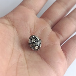 Op de afbeelding: Een zilveren en met diamanten bezette kraal met een traanvorm. De kraal is bezet met kleine, heldere diamanten en heeft een zwarte metalen band rond het midden.