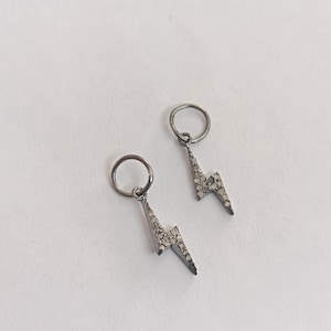 Può includere: Un paio di orecchini a forma di fulmine color argento, ciascuno ornato con piccoli abbellimenti scintillanti. Gli orecchini pendono da un piccolo cerchio, creando un effetto pendente. I gioielli sono posti su uno sfondo bianco.